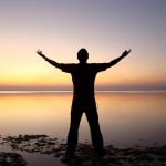 silhouette of man standing beside ocean during sunset